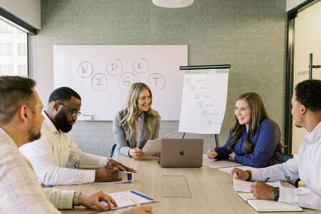 Business team in a conference room collaborating on a project, with two women leading the discussion in front of a whiteboard and flip chart, symbolizing teamwork, leadership development, and strategic planning.
