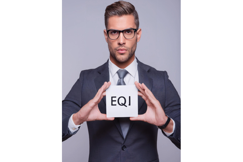 Confident businessman in a suit and glasses holding a white cube with the letters ‘EQI,’ representing emotional intelligence quotient.