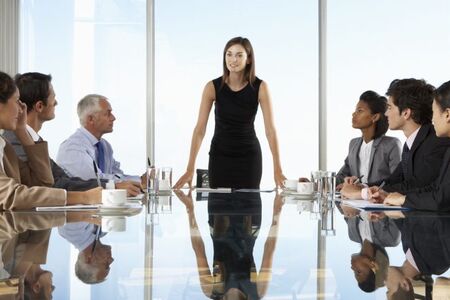 Businesswoman standing at the head of a conference table leading a meeting with a diverse group of professionals attentively listening.